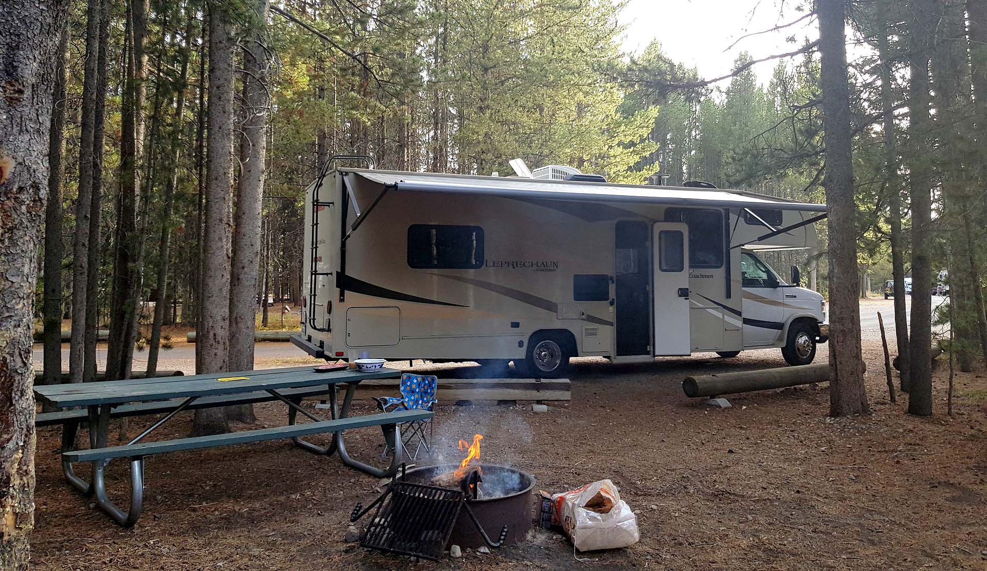 Class C RV parked in a campsite with fire in the fire pit. 
