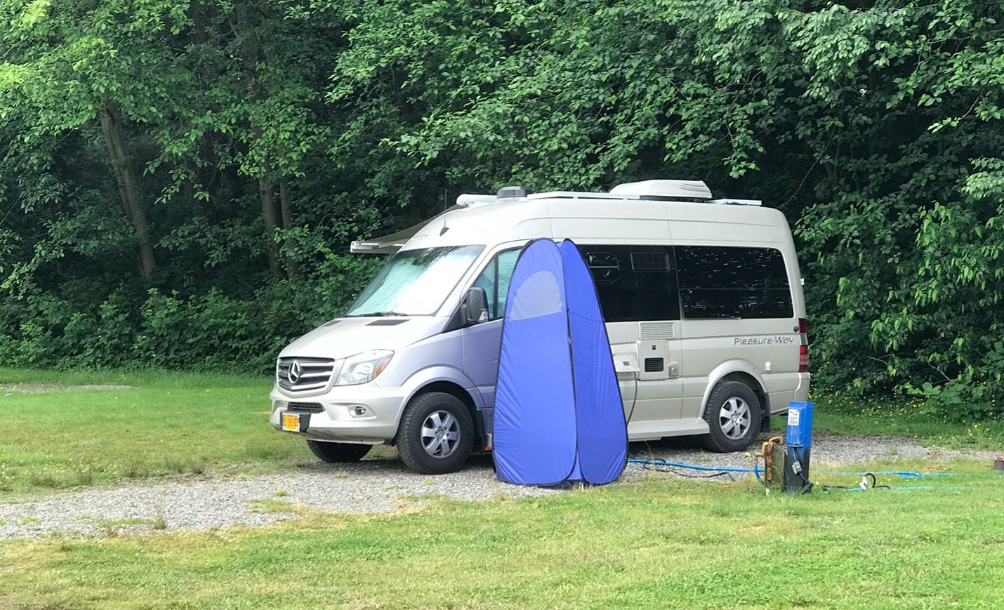 Silver RV van with a portable outdoor shower parked in a campground.